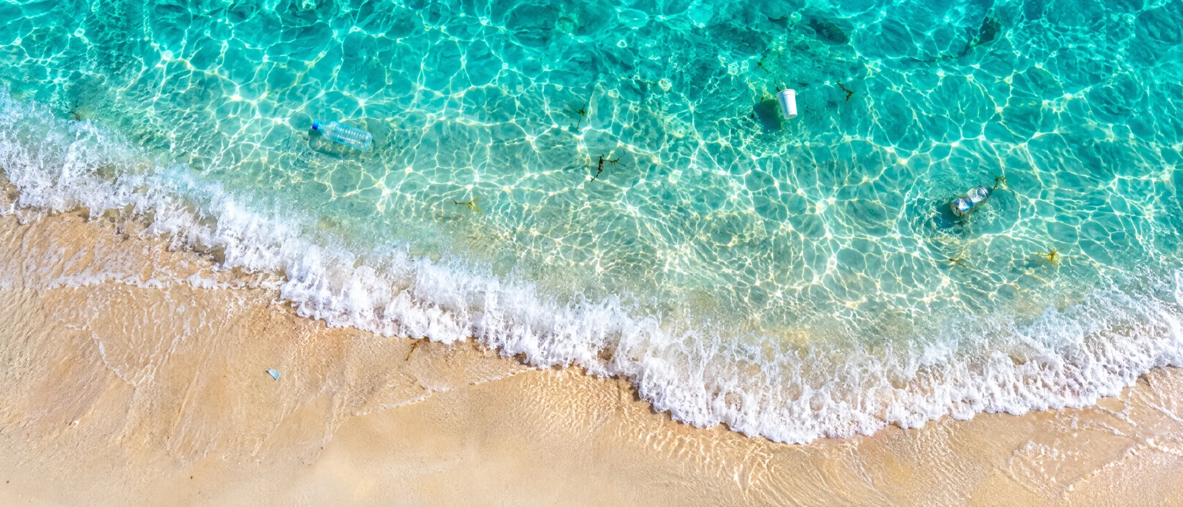 Aerial view straight down on turquoise shallows meeting pale sand, a few pieces of litter barely visible in the water